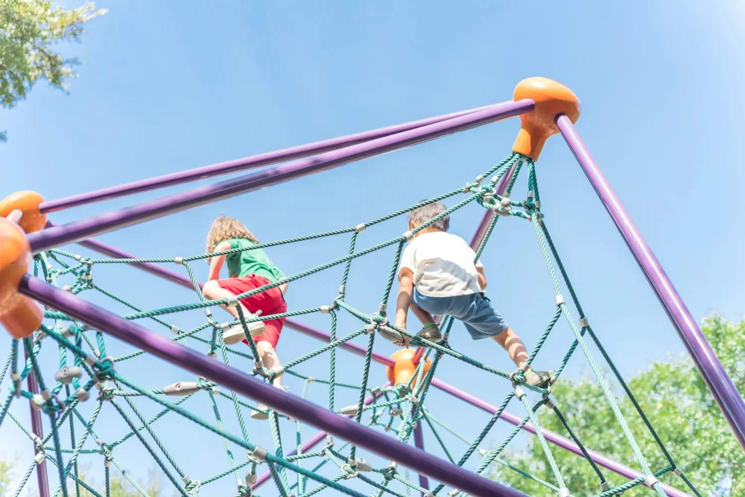 Two children climbing a rope playground structure against a blue sky
