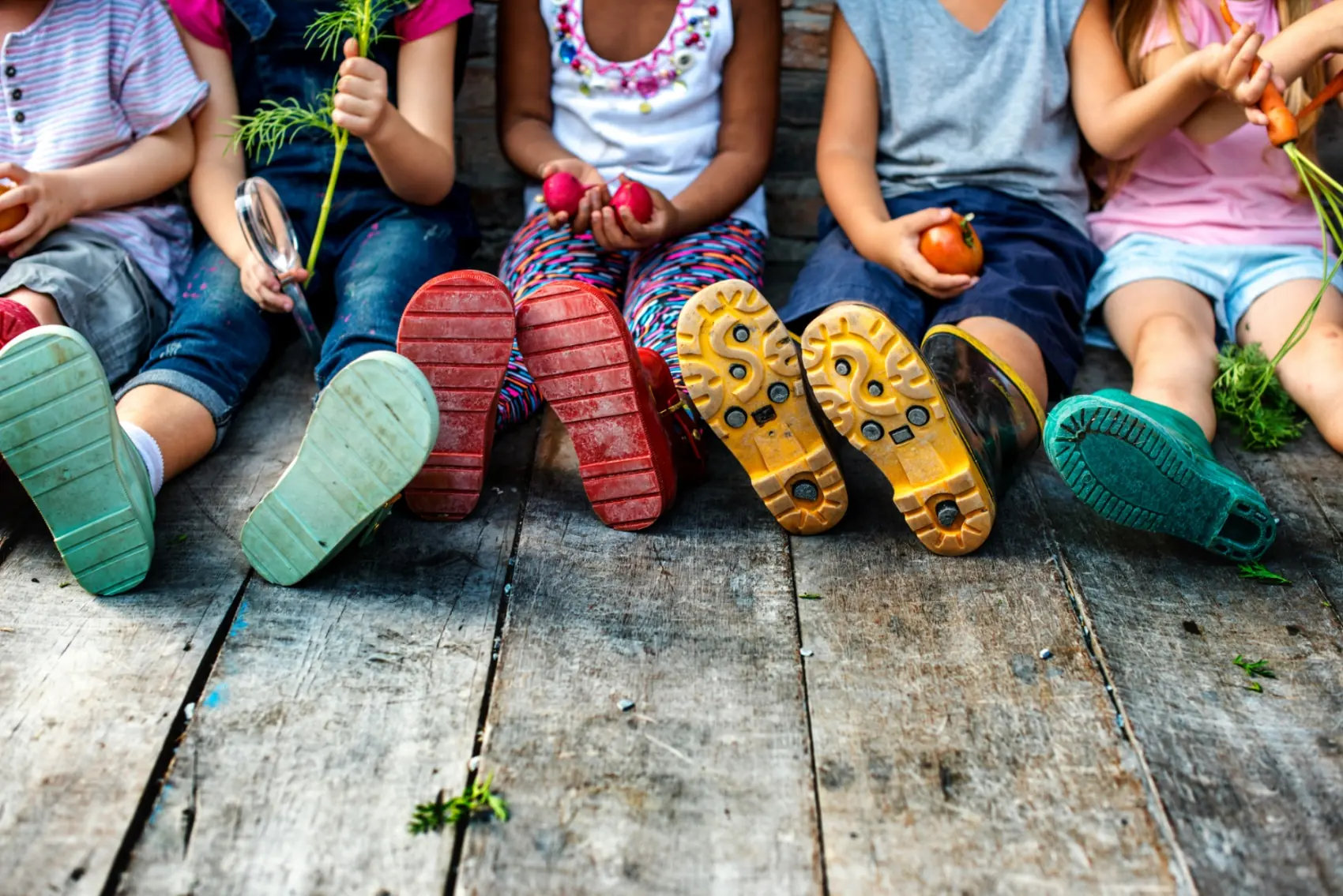 Children sitting in a row in rubber boots holding freshly picked vegetables